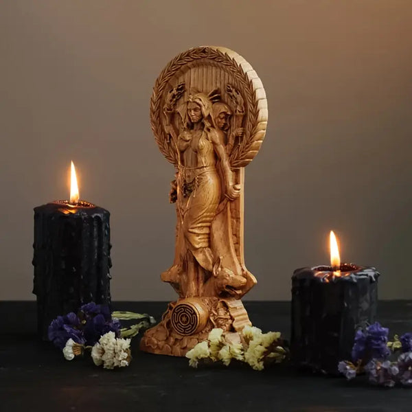 Wooden figurine of a woman with a wreath, flanked by two lit black candles on a dark surface. auraworks decor