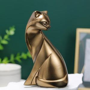 A metallic gold cat figurine sculpture sitting upright on a white surface against a dark green background with blurred decorative elements.