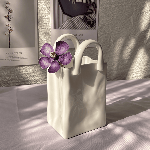 A cream-colored ceramic tote bag with dual top handles, styled with a purple flower on a light pink fabric surface with dappled sunlight and shadow, against a textured wall with framed prints.