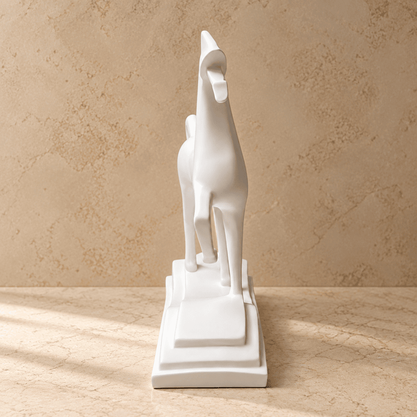 A minimalist white ceramic giraffe figurine standing on a tiered rectangular base, photographed against a warm beige textured wall and matching surface.