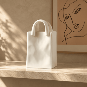 A matte white ceramic tote bag with integrated top handles sits on a light stone ledge in a warm, sunlit interior. A framed minimalist line drawing of a face hangs on the wall behind it.