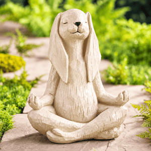 A beige stone-look rabbit garden statue sitting cross-legged in a meditative pose, photographed outdoors on a stone path with blurred green plants in the background.