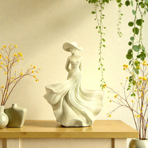 Decorative statue of a woman in a flowing dress on a wooden table with plants and vases. Ivory Serenity Goddess Sculpture