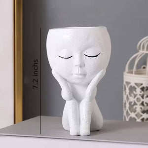 A white ceramic planter shaped like a seated face with closed eyes, shown on a tabletop with a gray wall behind it. A vertical measurement marker on the left reads 7.