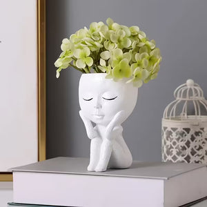 White ceramic vase shaped like a person with closed eyes and green flowers on a gray surface.