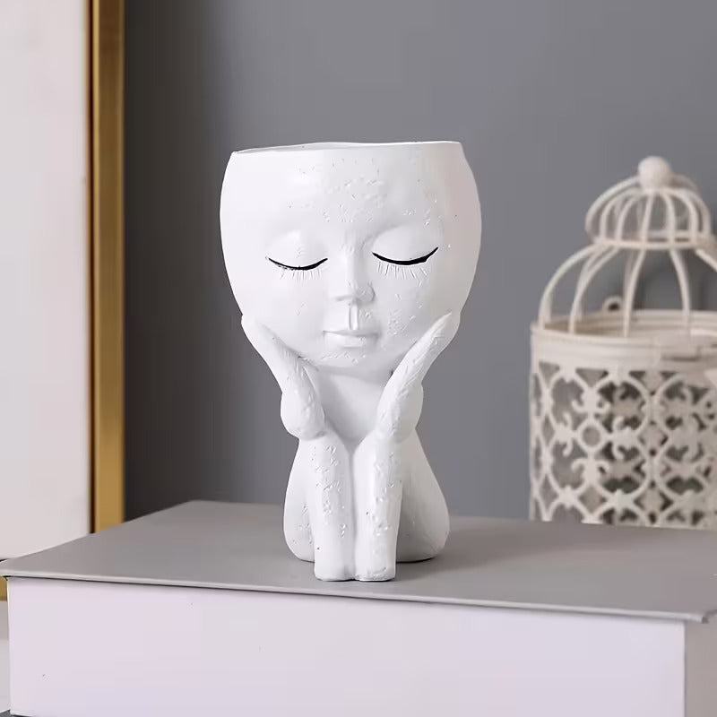 White ceramic vase shaped like a person with closed eyes and green flowers on a gray surface.