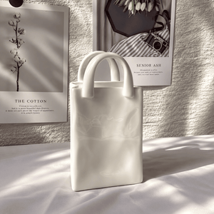 A matte white structured tote bag with two short top handles, standing on a white fabric surface in soft directional sunlight. Behind it are two framed black-and-white wall prints, one reading 'THE COTTON' and the other partially visible with 'SENIOR ASH'.