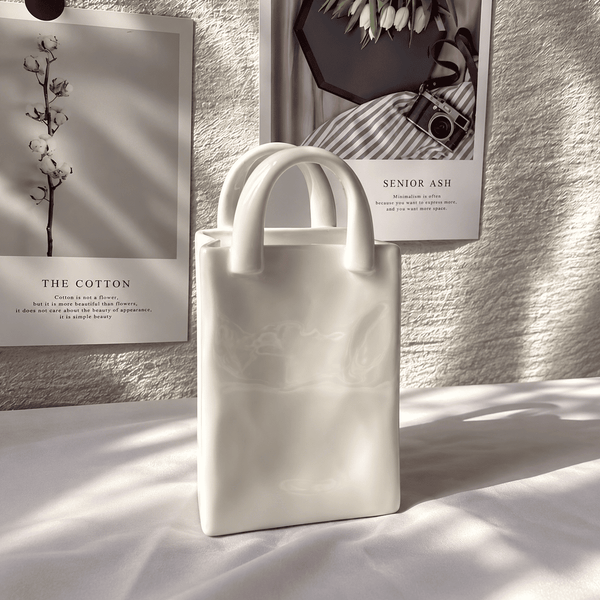 A matte white structured tote bag with two short top handles, standing on a white fabric surface in soft directional sunlight. Behind it are two framed black-and-white wall prints, one reading 'THE COTTON' and the other partially visible with 'SENIOR ASH'.