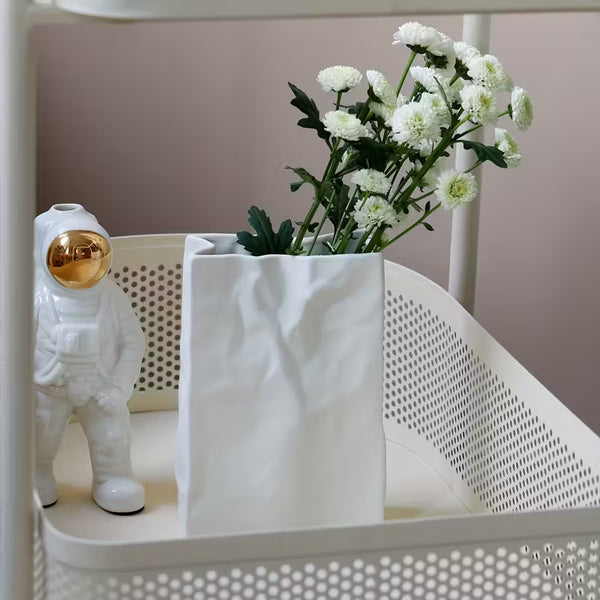 White vase with greenery and a small white figure in an astronaut suit on a perforated surface.