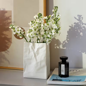 White vase with flowers on a surface next to a black bottle and open book.