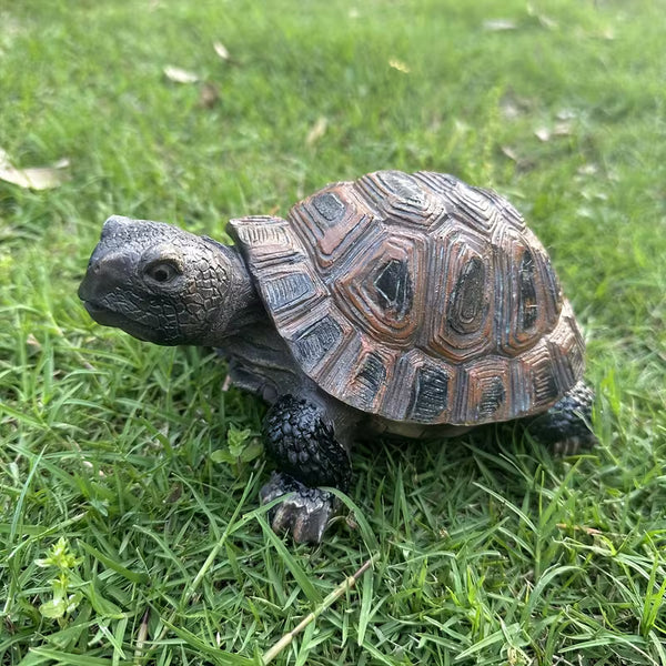A realistic tortoise statue with a patterned brown and gray shell and textured dark head and legs, standing on green grass.