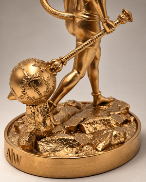 Close-up of a gold-toned figurative sculpture showing part of a human figure standing on a rocky base and holding a staff, with a round medallion and engraved base detail visible.