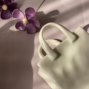 A glossy ceramic handbag-shaped object in a pale cream or off-white glaze, photographed on a soft pink surface with purple flowers and leaf shadows.