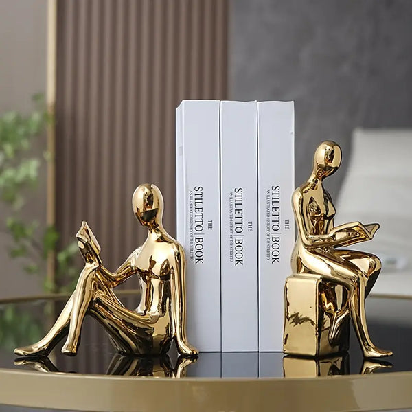 Gold human-shaped bookends on a reflective surface with books in the background. auraworks decor